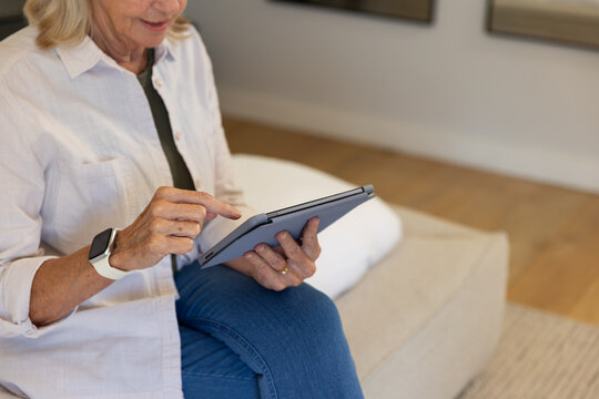 Senior woman sitting on ottoman tapping tablet at home wearing light shirt, jeans and smartwatch