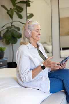 Senior woman sitting on bed wearing pink headset tapping tablet and checking smartwatch in bedroom
