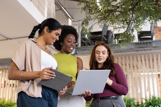 Diverse female coworkers reviewing report on silver laptop and tablet in office atrium