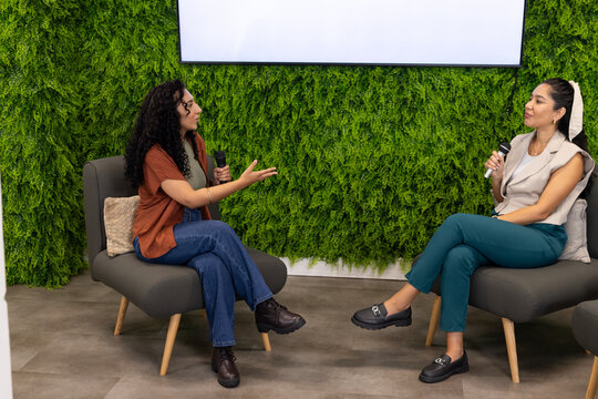 Two women sitting on gray lounge chairs in studio holding microphones, discussing by green wall