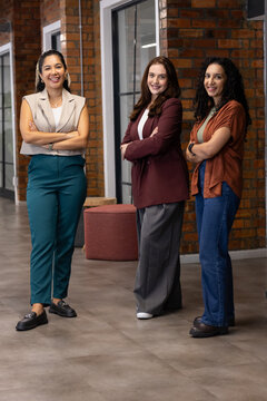 Three adult women standing with arms crossed in office corridor with exposed brick and glass doors