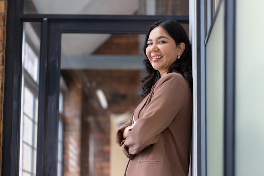 Woman leaning with back against glass partition in office corridor, wearing brown blazer