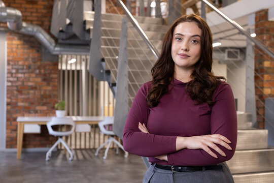 Woman in late 20s standing right of metal staircase in office, wearing maroon top, copy space