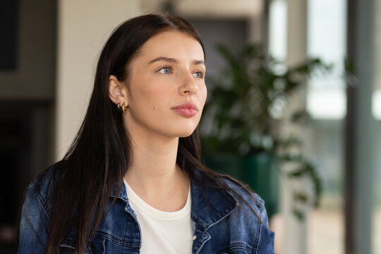 Woman in her 20s gazing upward right in modern room wearing denim jacket and gold hoops