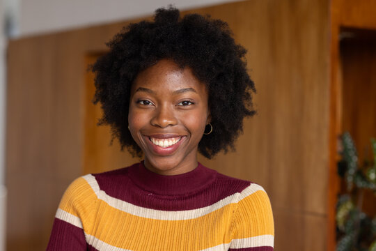 African woman smiling at camera wood wall doorway plant shelf wearing hoop earrings striped sweater