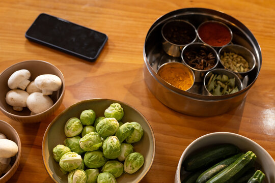 Bowl of Brussels sprouts sitting on wooden table with mushrooms, zucchini, spice tin nearby