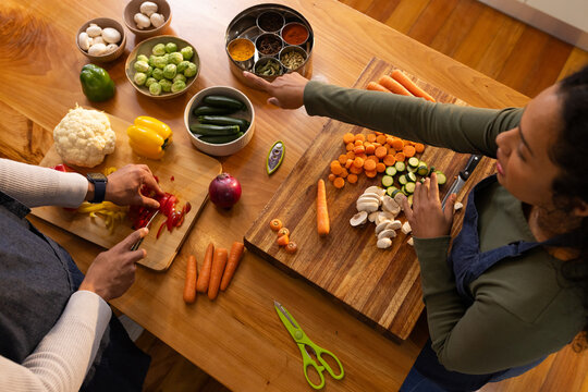 Couple wearing aprons chopping vegetables at kitchen table with cutting boards, reaching spice tin