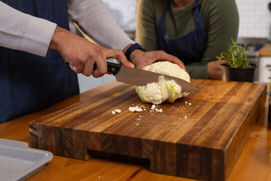 African American couple slicing cauliflower with chef's knife on butcher block in kitchen in aprons