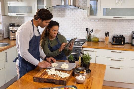 Couple wearing dark blue aprons preparing cheese on chopping board at kitchen island, using tablet