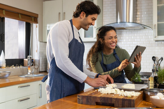 African American couple in blue aprons chopping on cutting board and reading tablet in home kitchen