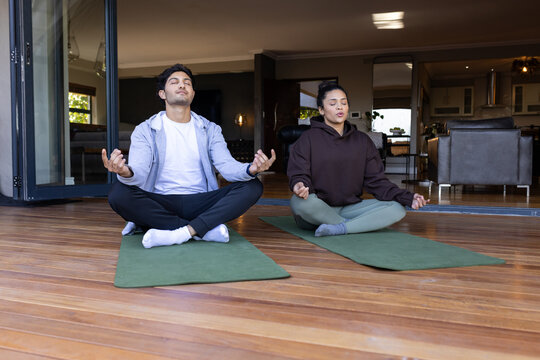 African American couple meditating wearing hoodies on green mats on wooden deck by folding doors