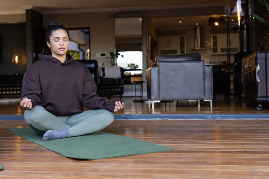 Adult African American woman sitting cross-legged on green mat on wood floor in hoodie and leggings
