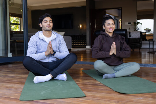 Couple meditating on green yoga mats at wooden deck outside sliding glass door, wearing hoodies