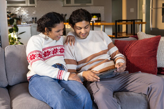 African American couple sitting on gray sofa with red pillows wearing knit sweaters using tablet