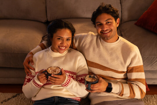 African American couple sitting on rug at home holding mugs, man wearing smartwatch, knit sweaters