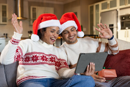 African American couple waving, holding tablet on sofa with wrapped gift, knit sweaters, Santa hats
