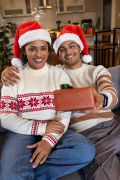 African American couple sitting on couch at home wearing Santa hats, man offering green bow gift