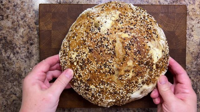 A close-up of female hands holding a round artisan sourdough loaf covered with crunchy sesame and flax seeds, gently pressing the golden crust to reveal its detailed texture.