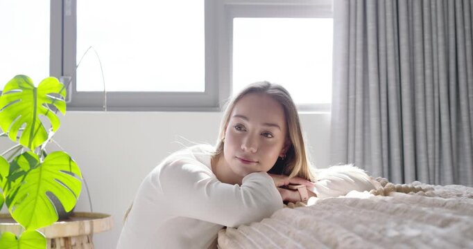 Female in her 20s leaning on textured coverlet by bright window, spotting camera and smiling