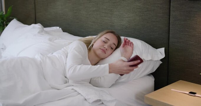 Woman in pajamas waking in bed, reaching for red smartphone to check time then settling