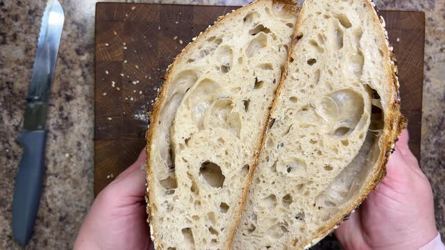 A top-down view of female hands holding two halves of freshly cut sourdough bread to show an open crumb with large air pockets, next to a knife on a wooden board.