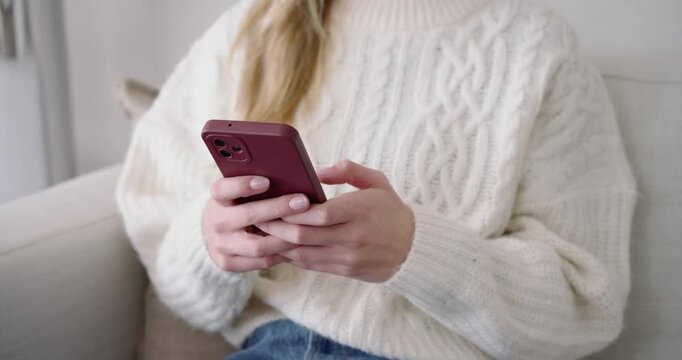 With thumbs resting on screen, woman is holding mauve phone and swiping to browse on sofa