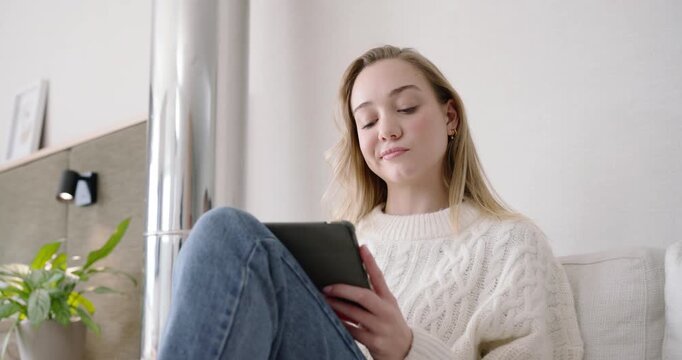Adult woman sitting on sofa reading tablet, reaching for credit card, entering payment, smiling