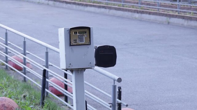 Metal access control terminal with keypad and adjacent black module mounted on pole beside railing; urban roadway and curb form backdrop