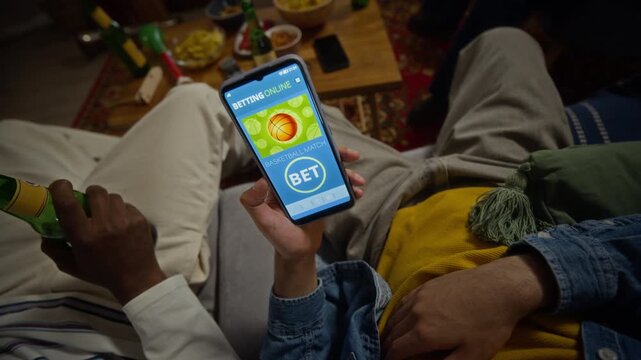 Multiethnic young adult men placing bets using mobile sports gambling app on smartphone at home. Clip suitable for betting platform advertising, online gaming services and digital fan campaigns