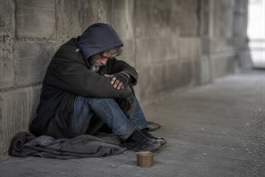 Sad elderly homeless man sitting on the street pavement with a begging cup, poverty and social exclusion concept