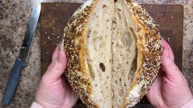 A close-up of a person holding two halves of freshly baked sourdough bread, showcasing the porous open crumb structure above a wooden cutting board with a knife.