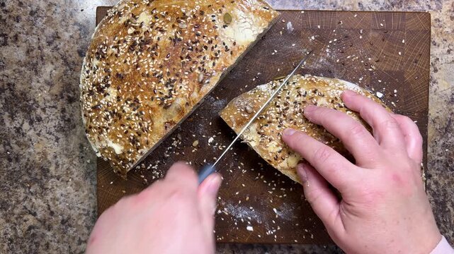 A close-up of a woman slicing freshly baked sourdough bread covered with crunchy sesame and flax seeds on a wooden cutting board in a kitchen.