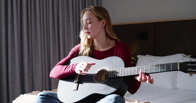 Woman starting by holding guitar in lap, strumming and adjusting frets, practicing on bed