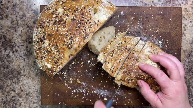 A close-up of a woman slicing freshly baked sourdough bread coated with sesame and flax seeds on a wooden cutting board in a kitchen.