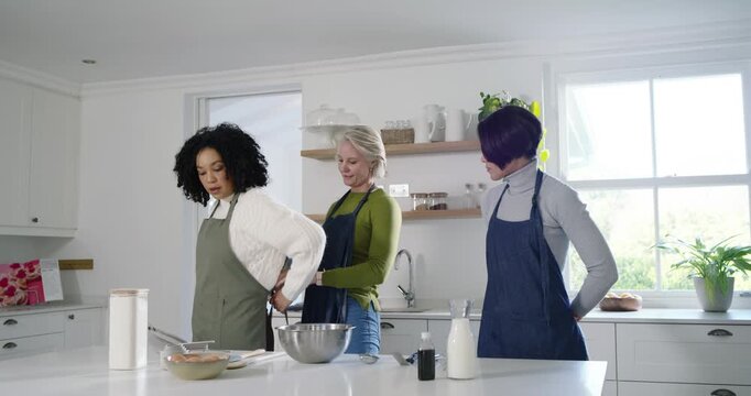 Female friends at kitchen island, center woman helping fasten aprons while left woman mixing batter