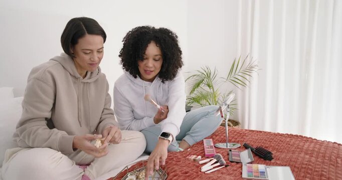 African American women sitting on bed testing makeup and jewelry, friend entering with dresses