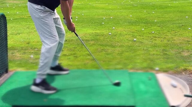 A golfer hitting a ball off a green practice mat with a club at a driving range, with the white ball flying into the distance and scattered golf balls visible in the background.