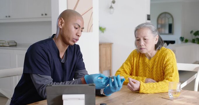 Diverse gloved caregiver testing Asian senior's finger with glucometer, checking blood sugar