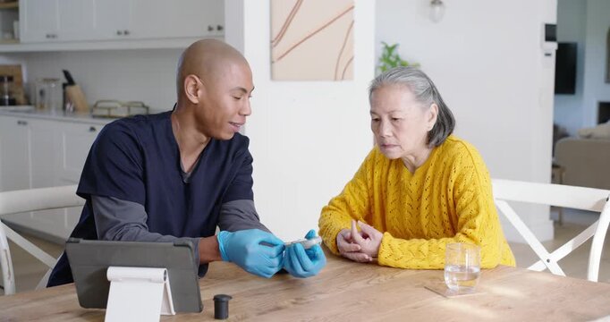 African American caregiver checking Senior Asian woman's glucose during check in kitchen with meter