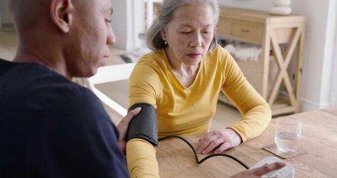 Senior Asian woman sitting at home table African American aide fitting cuff checking BP