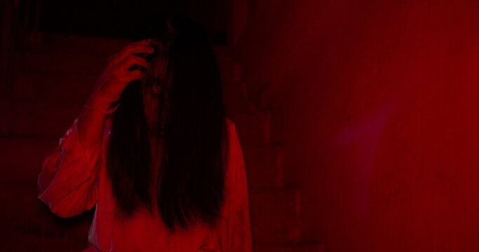 A terrifying female ghost emerges from the shadows of a dark, red-lit basement, creating a suspenseful and scary scene perfect for a Halloween horror movie trailer.