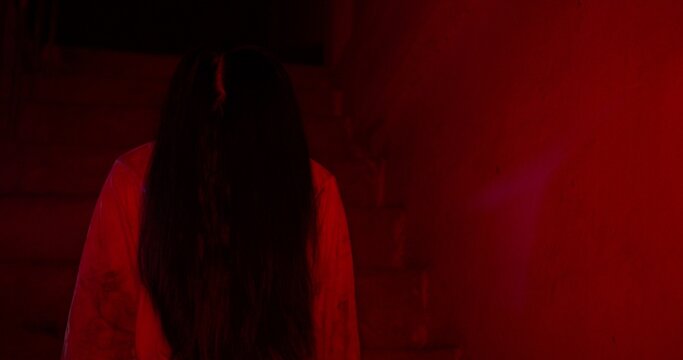 A terrifying female ghost emerges from the shadows of a dark, red-lit basement, creating a suspenseful and scary scene perfect for a Halloween horror movie trailer.
