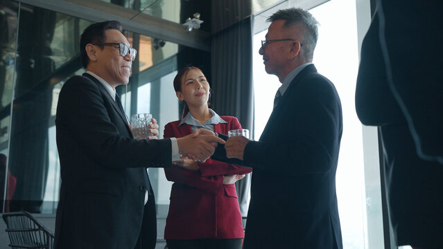 Two businessmen in suits shake hands while holding drinks, with a businesswoman in a red suit watching smilingly at a corporate event
