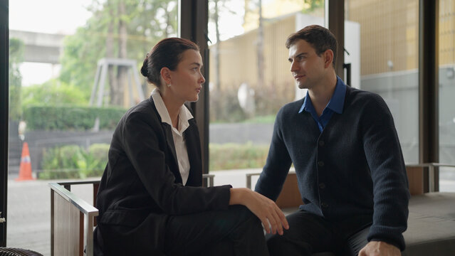 A woman in a black suit and a man in a blue cardigan sit near large windows, looking at each other during a conversation