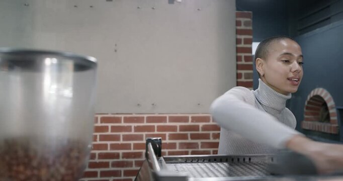 African woman in turtleneck checking espresso machine, wiping cloth behind cafe counter by hopper