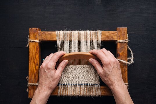 Hands of an artisan weaving handmade wool textile with a wooden comb on a small frame loom over a dark background