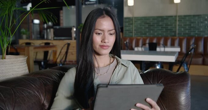 Mid adult Asian woman in green shirt browsing tablet in folio in cafe, tapping, swiping, smiling