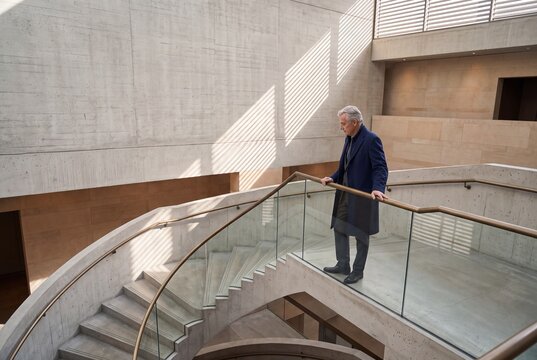 Senior businessman in a dark coat standing thoughtfully on a modern concrete staircase inside a contemporary architectural building