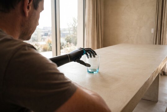 Man with an advanced bionic prosthetic arm reaching for a glass on a table in a modern home
