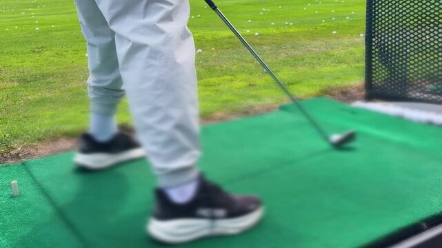 A golfer hitting a ball off a green practice mat with a club at a driving range, with the white ball flying into the distance, scattered golf balls, and a training net in the background.